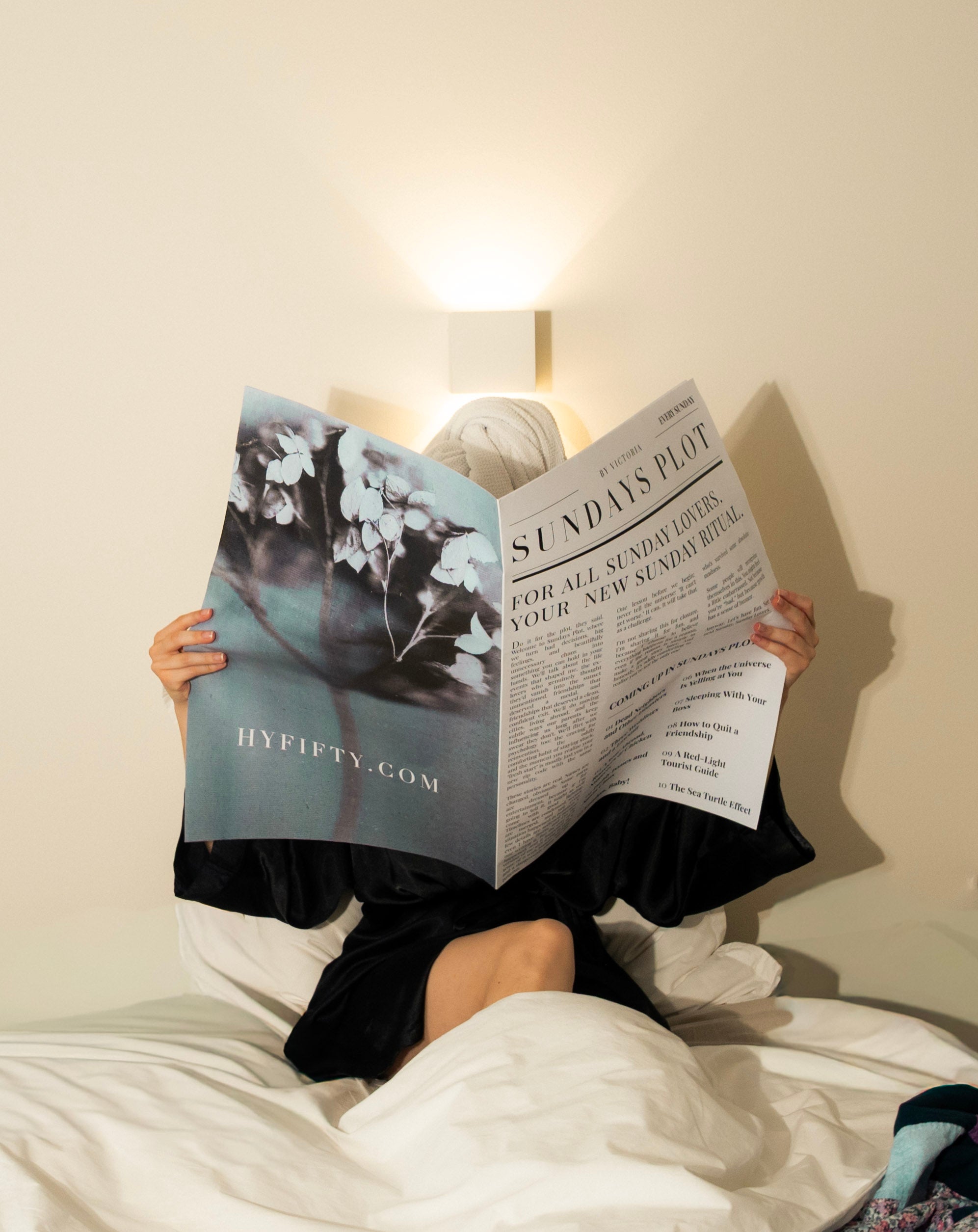 Editorial scene of a woman in a black silk robe lying on the bed, feet resting under the sheets, reading a newspaper.