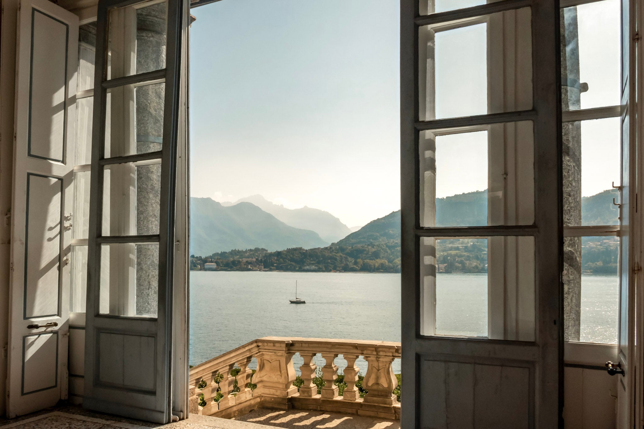 Open doors frame a sunlit lake view with mountains, a balcony and a small sailboat in the distance.