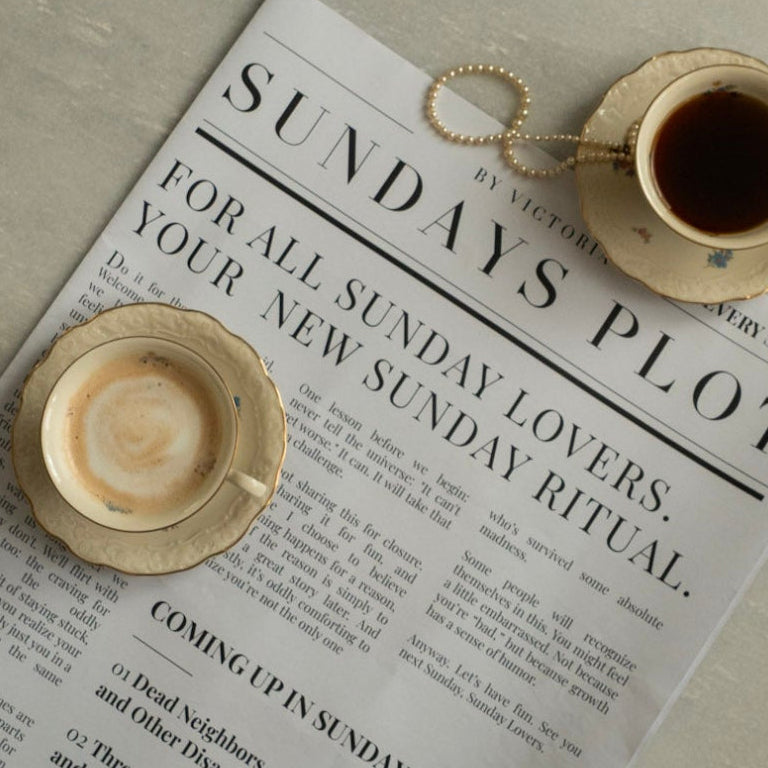 Sundays Plot newspaper on a round table with coffee cups, a pearl necklace and soft morning light.